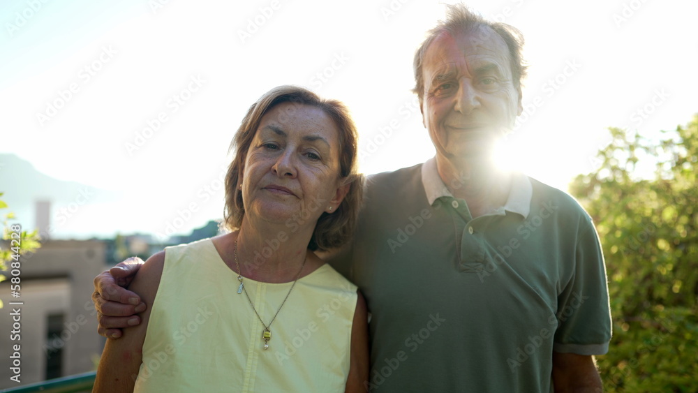 Naklejka premium Portrait of a senior couple standing outdoors during sunset posing for camera. An older husband with arm around wife