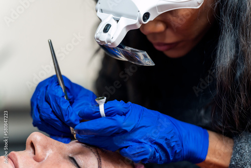 Crop black concentrated cosmetologist with black hair wearing magnification goggles while doing permanent makeup on mature clients eyebrows in beauty salon