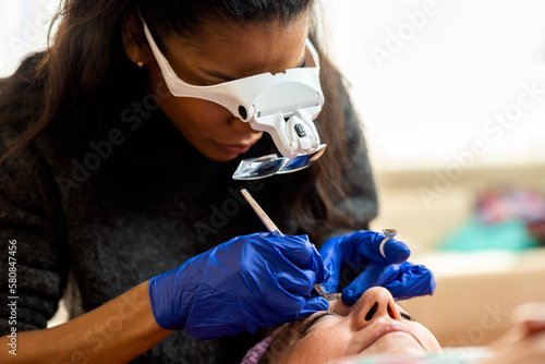 Crop black professional cosmetician in gloves and magnification goggles for microblading applying permanent makeup with pen in beauty clinic