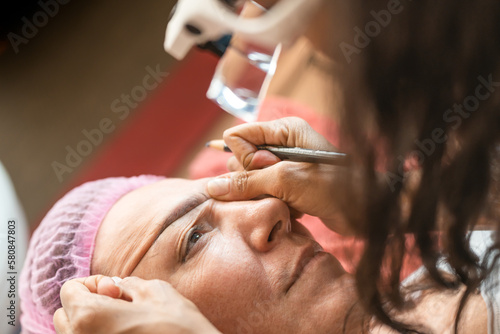High angle of crop anonymous female cosmetologist doing brow shape correction with thread on client in beauty salon