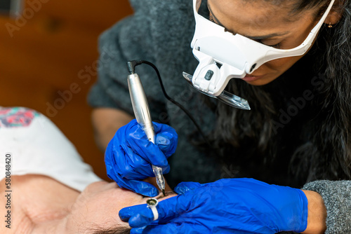 Crop professional black cosmetologist wearing microblading magnification goggles while doing permanent eyebrows tattoo makeup with handheld tool