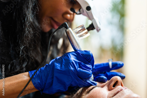 Side view of crop African American female cosmetologist in magnification goggles and blue gloves with tattoo ink ring applying permanent eyebrows makeup with needles