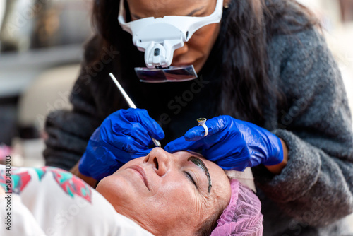 Crop microblading master in magnification goggles applying permanent makeup on eyebrows of female client with pen during beauty session in modern salon