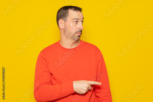 Bearded hispanic man wearing a red sweater pointing to the side with the index finger isolated on yellow studio background