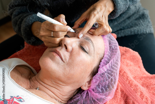 From above of crop anonymous female cosmetologist preparing mature woman for eyebrow permanent makeup procedure in beauty salon