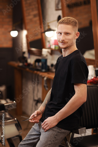 Stylish barber with beard and mustache man in casual work clothes holds professional scissors for haircut stands in hair salon. Confident hairdresser stands near workplace in barbershop. Beaty salon