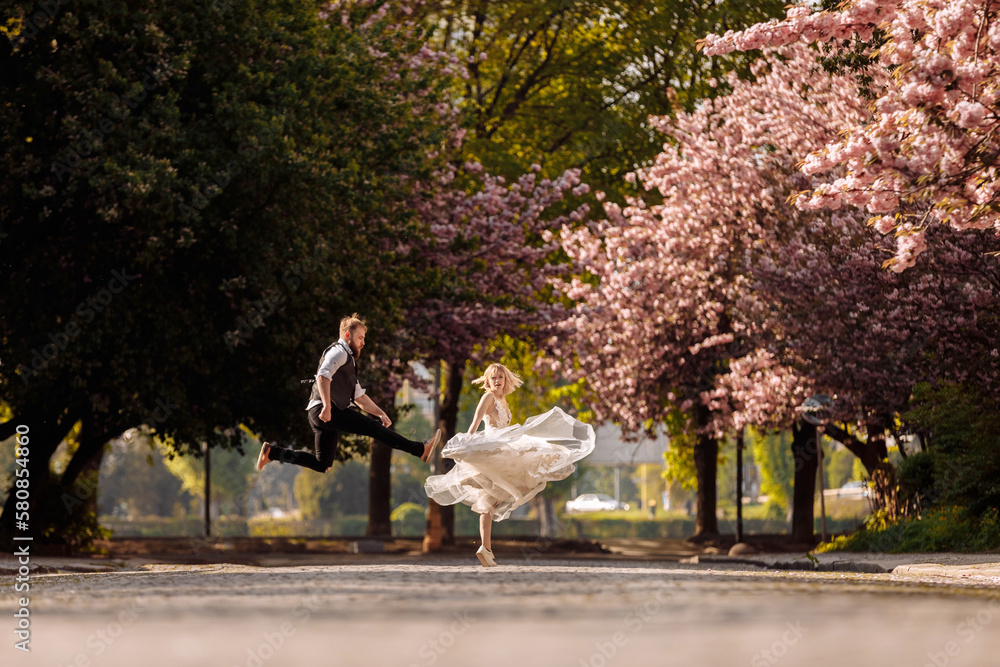 Obraz premium Beautiful, cheerful and lively newlyweds, groom and bride are hugging near the blooming pink cherry blossom. Wedding portrait of a close-up of a smiling bearded groom and a cute bride.