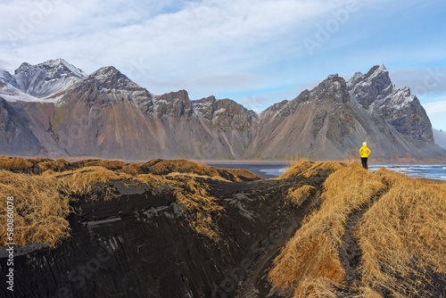 femme en ciré jaune au bord d'une plage de sable noir en Islande avec des montagne enneigés 