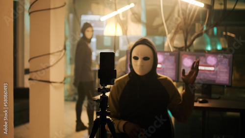 Skilled cyber criminal with white mask recording live ransomware video on dark web, making threats after stealing passwords. Masked hacker acting mysterious on camera at night, harassment.