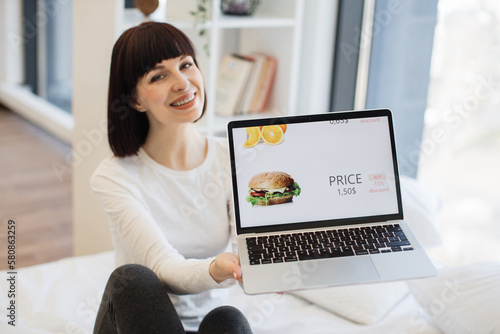Smiling dark haired woman in casual attire sitting on bed at home and using modern laptop for online shopping. Focus on computer screen with tasty burger selling with 15 percent discount.