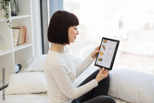 Attractive caucasian woman in casual attire sitting on bed with digital table in hands and scrolling online shop with fast food. Concept of grocery, shopping and gadgets.