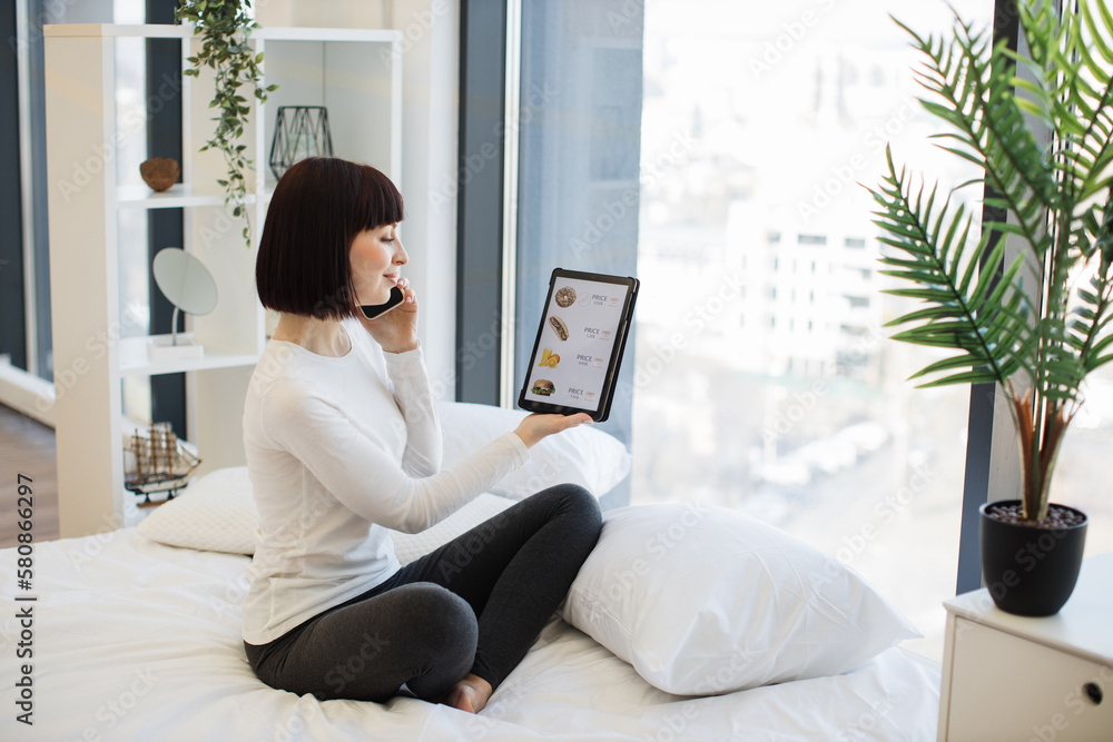 Casually dressed woman holding digital tablet and having mobile conversation while sitting on cozy white bed. Happy lady staying at home and ordering food for lunch in online store with discount. Casually dressed woman holding digital tablet and having mobile conversation while sitting on cozy white bed. Happy lady staying at home and ordering food for lunch in online store with discount.