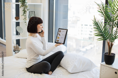 Casually dressed woman holding digital tablet and having mobile conversation while sitting on cozy white bed. Happy lady staying at home and ordering food for lunch in online store with discount.