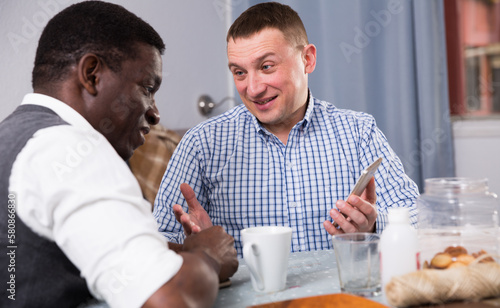 Interracial two male friends using phone and talking in home interior