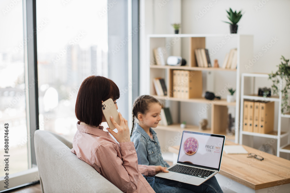 Back view of woman ordering pizza via smartphone while sitting with little girl on sofa. Pleasant mother and cute daughter spending quality time together while buying takeaway lunch with laptop. Back view of woman ordering pizza via smartphone while sitting with little girl on sofa. Pleasant mother and cute daughter spending quality time together while buying takeaway lunch with laptop.
