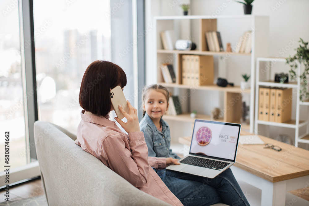Back view of woman ordering pizza via smartphone while sitting with little girl on sofa. Pleasant mother and cute daughter spending quality time together while buying takeaway lunch with laptop. Back view of woman ordering pizza via smartphone while sitting with little girl on sofa. Pleasant mother and cute daughter spending quality time together while buying takeaway lunch with laptop.