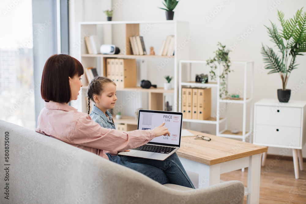 Caring mother with little daughter browsing ecommerce site for searching discount of new trendy apparel. Young woman and pretty girl sitting together on comfy couch and using modern wireless laptop. Caring mother with little daughter browsing ecommerce site for searching discount of new trendy apparel. Young woman and pretty girl sitting together on comfy couch and using modern wireless laptop.