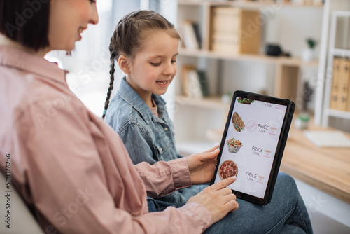 Close up of smiling shoppers choosing fast food at discount on delivery webpage while relaxing at home. Serene woman and girl spending quality time together while buying precooked meals via tablet.
