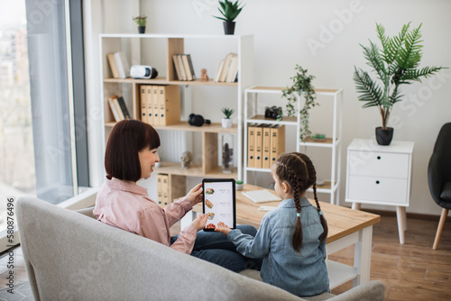 Happy mother and daughter resting on couch and pointing with their hands at tablet screen where junk food service online app being shown. Cheerful female shoppers making use of e-commerce.