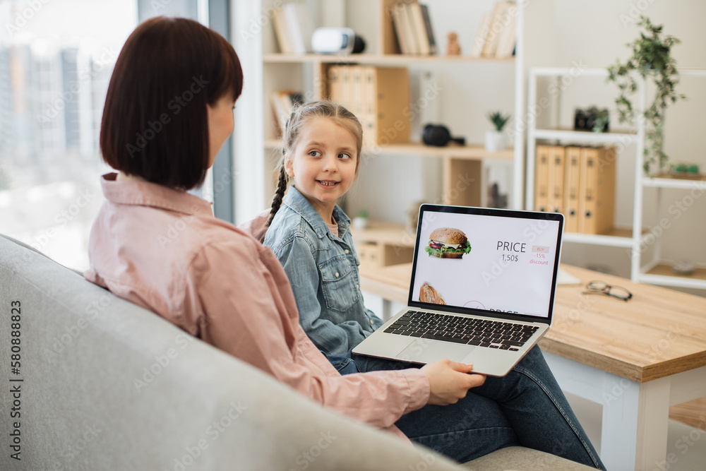 Attractive dark haired woman sitting on couch with her lovely daughter and choosing fast food with discount on website. Caucasian family of two using modern laptop for ordering takeaway lunch. Attractive dark haired woman sitting on couch with her lovely daughter and choosing fast food with discount on website. Caucasian family of two using modern laptop for ordering takeaway lunch.