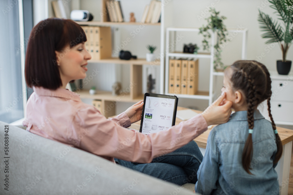 Blurred foreground of attractive mother in apartment touching daughter's cheek while holding electonic device in hand. Focus on digital tablet with virtual cosmetics store website offering discounts. Blurred foreground of attractive mother in apartment touching daughter's cheek while holding electonic device in hand. Focus on digital tablet with virtual cosmetics store website offering discounts.