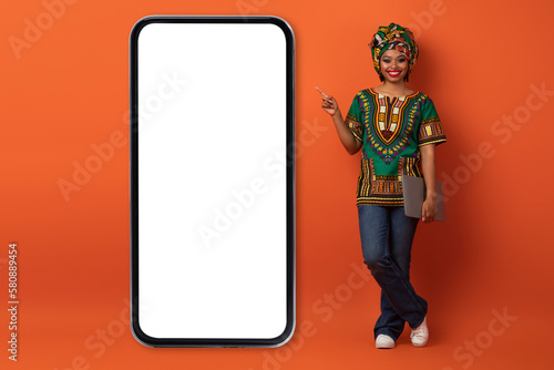 Happy african woman with laptop pointing at phone screen © Prostock-studio
