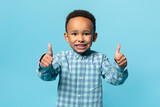 I like it. Happy african american adorable boy showing thumbs up and widely smiling, posing over blue background