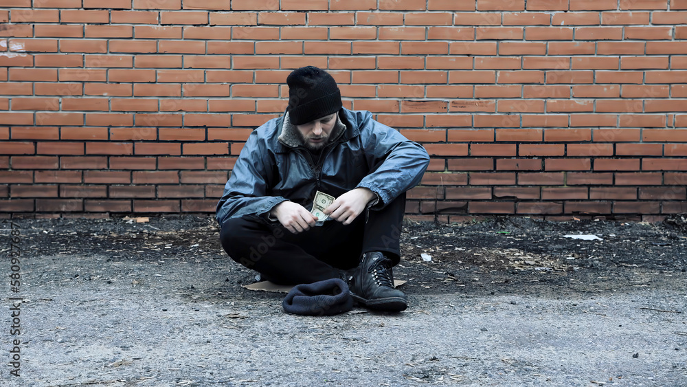 Homeless beggar holds cash money in his hands. Refugee needs help. Tramp sits at brick wall wearing in dirty clothes, hat cap. Below poverty line. Fugitive. Illegal immigrant. Drunkenness, alcoholism