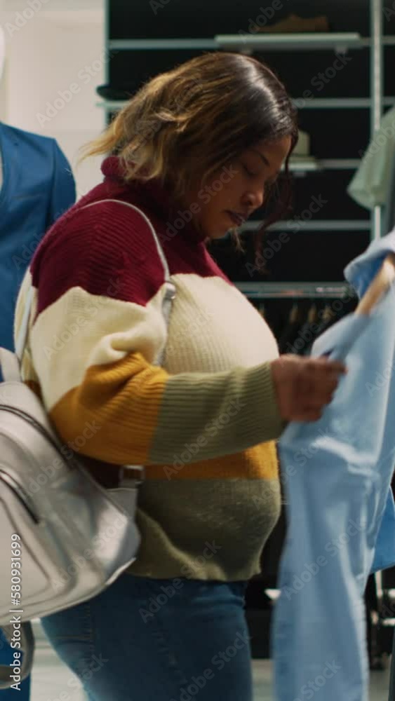 Vertical video: African american woman taking formal wear from hangers to increase wardrobe in clothing store. Young client trying to buy new clothes collection, analyzing fabric in boutique. Handheld Vertical video: African american woman taking formal wear from hangers to increase wardrobe in clothing store. Young client trying to buy new clothes collection, analyzing fabric in boutique. Handheld