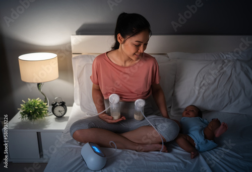 mother using breast pump machine to pumping milk while talking with her newborn baby on bed at night