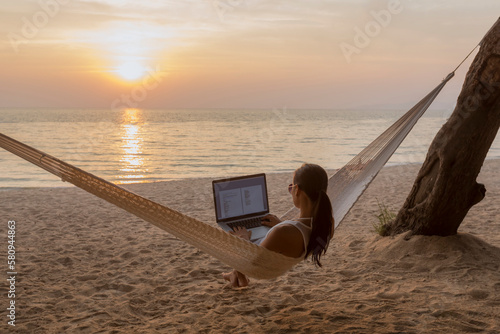 Woman using laptop computer while sitting on hammock at beach photo