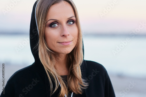 Beauty casual look. Young woman in black hood over the dusk sky and sea.