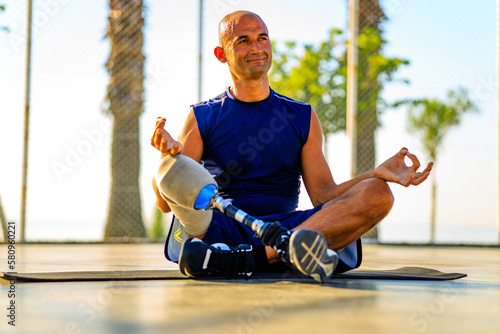 man with prosthesis leg practicing yoga sitting in lotus pose on yoga mat meditating outdoors in sun lights