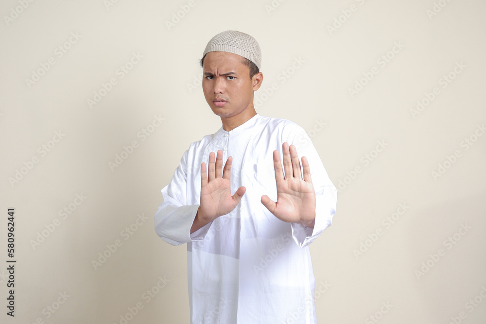 Portrait of attractive Asian muslim man in white shirt with skullcap forming a hand gesture to avoid something or bad things. Isolated image on grey background Portrait of attractive Asian muslim man in white shirt with skullcap forming a hand gesture to avoid something or bad things. Isolated image on grey background