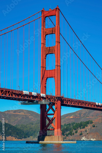 Fototapeta Naklejka Na Ścianę i Meble -  Majestic golden gate bridge with suspension wires and red girders on exterior near blue clear sky and ocean mountains
