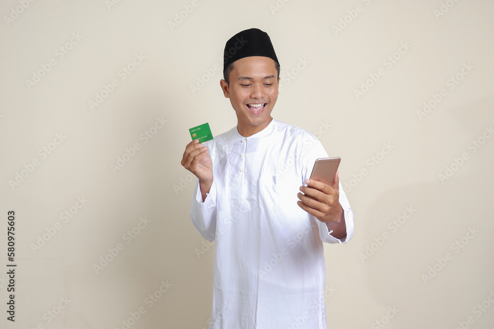 Portrait of attractive Asian muslim man in white shirt with skullcap holding a mobile phone and presenting credit card. Isolated image on gray background Portrait of attractive Asian muslim man in white shirt with skullcap holding a mobile phone and presenting credit card. Isolated image on gray background