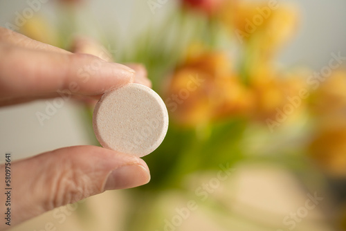 round large tablet, vitamin in the girl's hand, against the background of yellow tulips, indoors. Flu and cold season, new virus strain. Lack of vitamins in spring and winter, uncontrolled medication