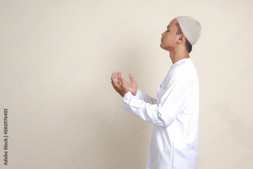 Portrait of attractive Asian muslim man in white shirt with skullcap praying earnestly with his hands raised. Isolated image on gray background Portrait of attractive Asian muslim man in white shirt with skullcap praying earnestly with his hands raised. Isolated image on gray background
