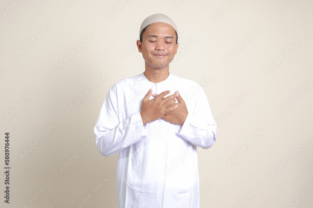 Portrait of attractive Asian muslim man in white shirt with skullcap placing hand on heart, feeling very grateful. Isolated image on gray background