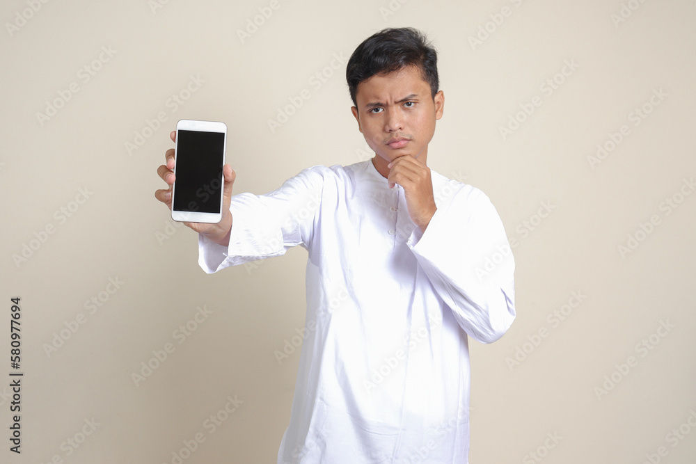 Portrait of attractive Asian muslim man in white shirt showing and presenting blank screen mobile phone. Advertising concept. Isolated image on gray Portrait of attractive Asian muslim man in white shirt showing and presenting blank screen mobile phone. Advertising concept. Isolated image on gray