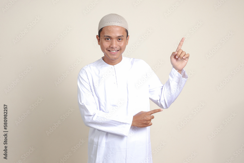 Portrait of attractive Asian muslim man in white shirt with skullcap showing product and pointing with his hand and finger to the side. Advertising concept. Isolated image on gray background Portrait of attractive Asian muslim man in white shirt with skullcap showing product and pointing with his hand and finger to the side. Advertising concept. Isolated image on gray background