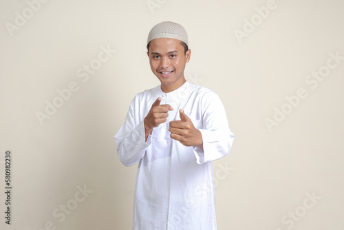 Portrait of attractive Asian muslim man in white shirt with skullcap showing product and pointing with his hand and finger to the side. Advertising concept. Isolated image on gray background