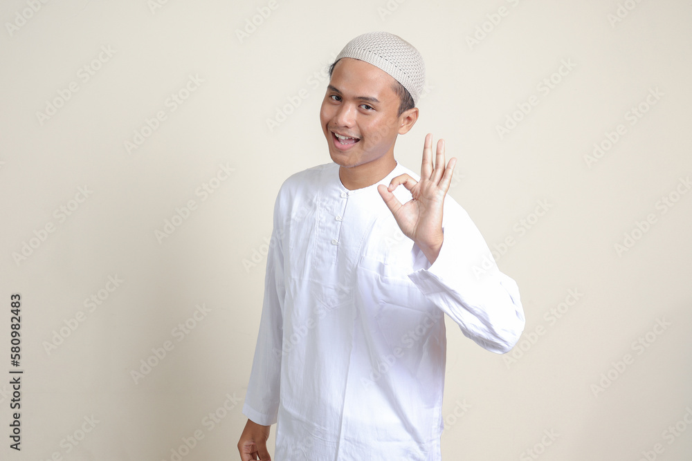 Portrait of attractive Asian muslim man in white shirt with skullcap showing ok hand gesture and smiling looking at camera. Advertising concept. Isolated image on gray background