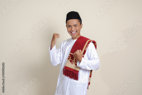 Portrait of attractive Asian muslim man in white shirt raising his fist, celebrating success. Isolated image on gray background