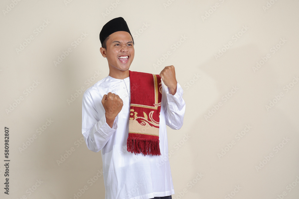 Portrait of attractive Asian muslim man in white shirt raising his fist, celebrating success. Isolated image on gray background Portrait of attractive Asian muslim man in white shirt raising his fist, celebrating success. Isolated image on gray background