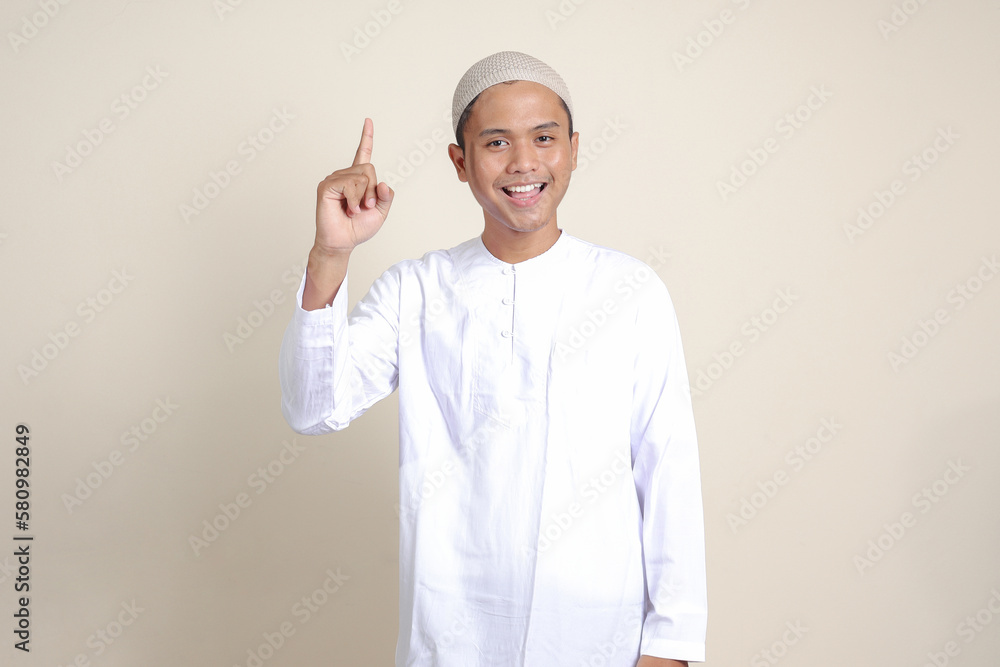 Portrait of attractive Asian muslim man in white shirt pointing up his forefinger and reminding something to do. Advertising concept. Isolated image on gray background Portrait of attractive Asian muslim man in white shirt pointing up his forefinger and reminding something to do. Advertising concept. Isolated image on gray background