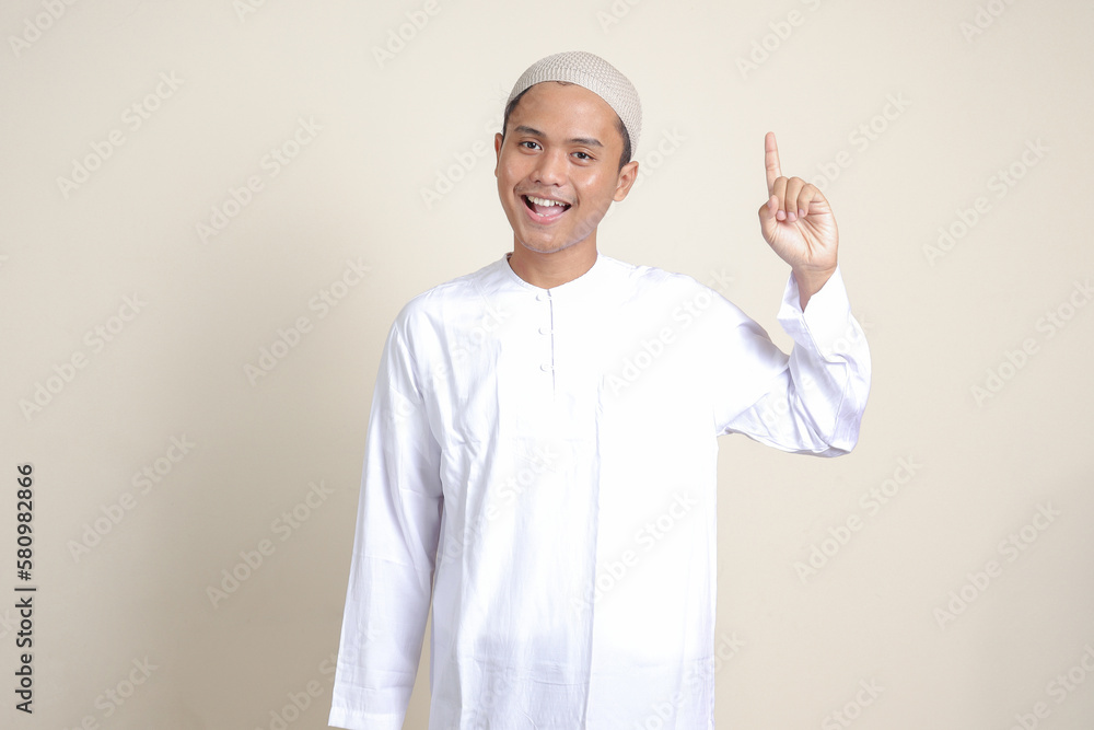 Portrait of attractive Asian muslim man in white shirt pointing up his forefinger and reminding something to do. Advertising concept. Isolated image on gray background Portrait of attractive Asian muslim man in white shirt pointing up his forefinger and reminding something to do. Advertising concept. Isolated image on gray background