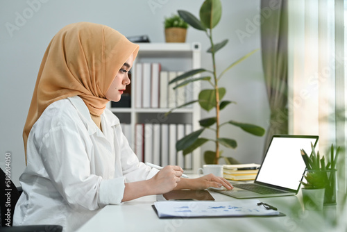 Concentrated muslim woman economist analyzing financial market on laptop computer at work station