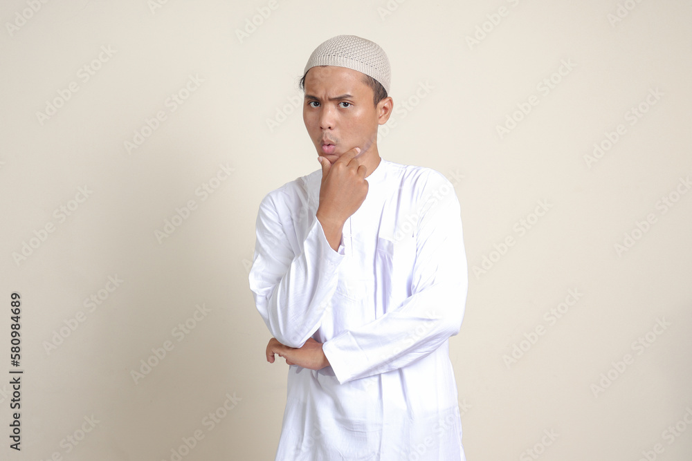 Portrait of attractive Asian muslim man in white shirt thinking about question with hand on chin. Isolated image on gray background Portrait of attractive Asian muslim man in white shirt thinking about question with hand on chin. Isolated image on gray background