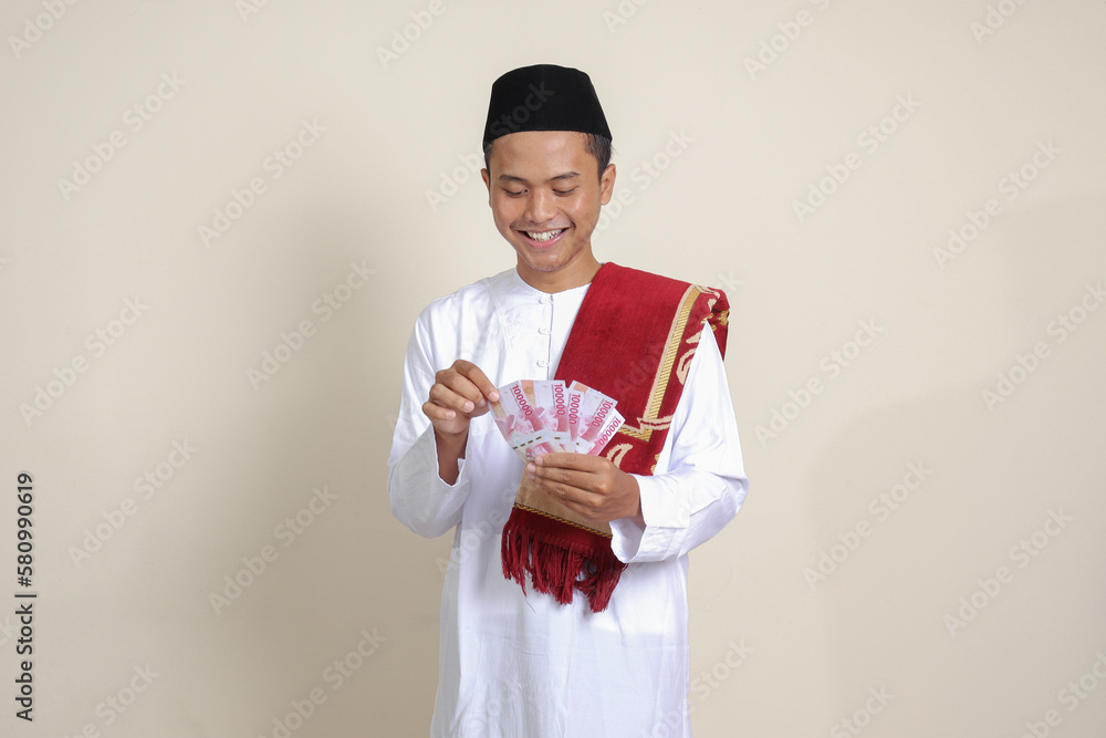 Portrait of attractive Asian muslim man in white shirt showing one hundred thousand rupiah. Financial and savings concept. Isolated image on gray background Portrait of attractive Asian muslim man in white shirt showing one hundred thousand rupiah. Financial and savings concept. Isolated image on gray background